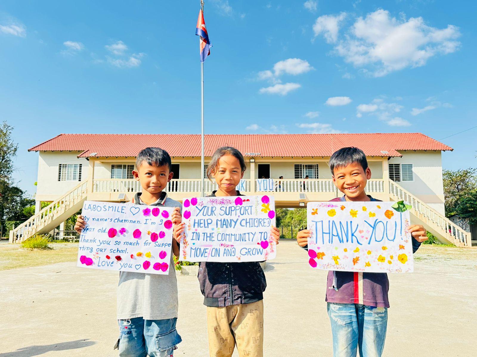 Trois enfants devant l'école avec des pancartes faites main remerciant LaboSmile
